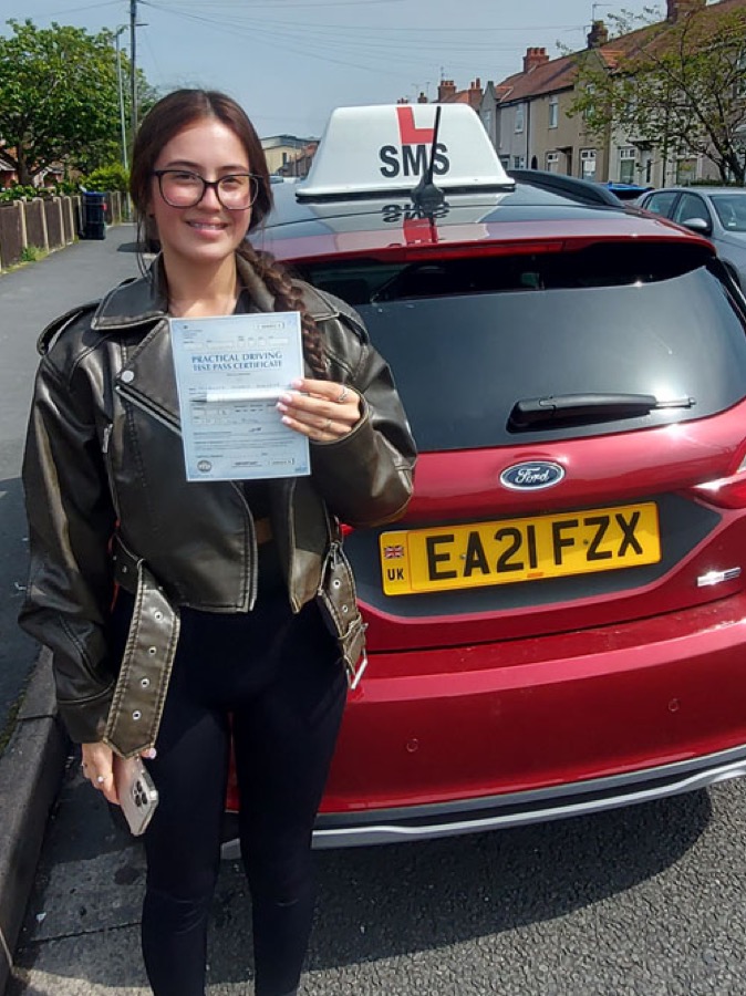 An SMS Driving School pupil holding their practical driving test pass certificate beside the school's red Ford Fiesta with its SMS roof sign